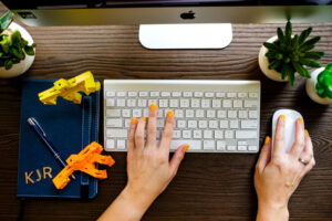A couple of hands on keyboard of a computer working. with a KJR notebook next to her hands.